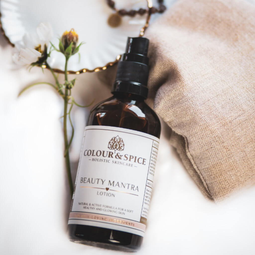 Op zachte ondergrond Beauty mantra lotion 100 ml huidverzorging olie in bruinglazen flesje van natuurlijk holistisch Beautymerk Colour and Spice met bloemtakje en authentieke luxe materialen als linnen porselein hout wellness